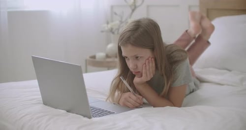 Niña aburrida está haciendo la tarea en el cuaderno, mirando en la pantalla del portátil durante las lecciones en línea