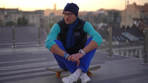 Positive Man Sitting on Skateboard Singing Dancing with Urban City at Sunset at Background
