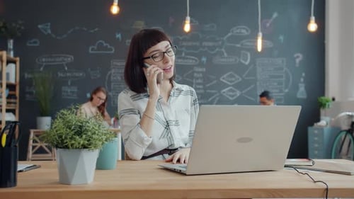 Female Office Worker Talking on Mobile Phone Using Laptop Working in Modern Workplace