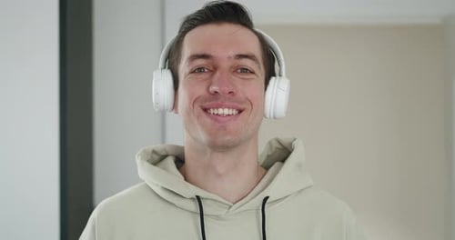 Portrait of a Stylish Man in White Headphones Listening to Music and Looking at the Camera Indoors