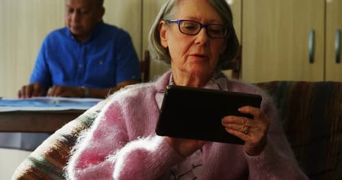 Senior Woman Using Tablet, Man Reads in Background
