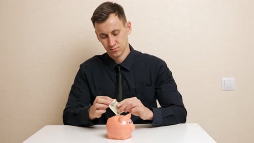 Man Takes Dollar Banknotes Out of Shirt Breastpocket and Inserts Into Small Pink Piggy Bank Slot