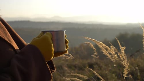 Person Holding Metal Mug in Rural Landscape at Sunrise