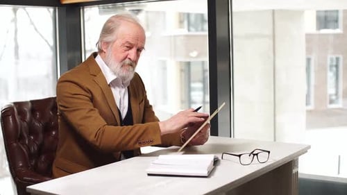 Senior Man Writes in Notebook at Desk with Tablet
