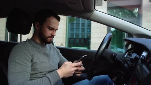 Serious bearded young man sitting in car and typing online message on cell phone, side view.