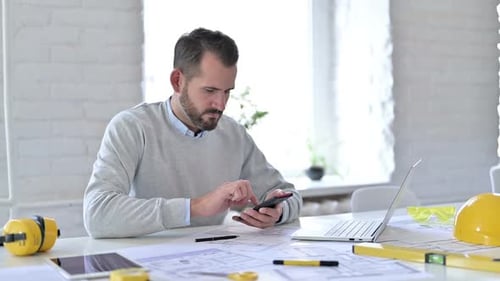 Engineer uses Smart Phone at Desk with Blueprints