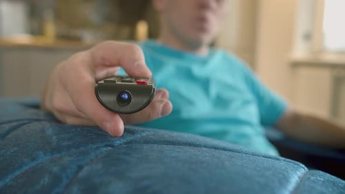 Nahaufnahme der Hand eines Mannes mit einer Fernsehfernbedienung, der zu Hause auf der Couch sitzt