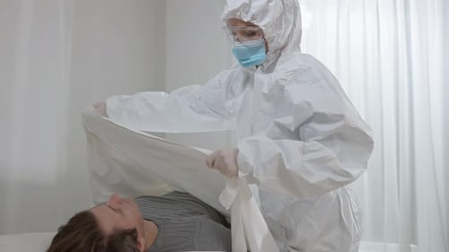 Infirmière en costume de protection, lunettes et masque facial couvrant un homme mort avec un drap blanc