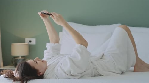 Woman in Robe Using Phone While Lying on Bed