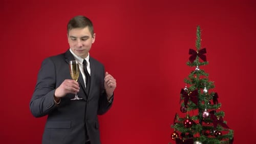 Jovem dançando com uma taça de champanhe perto da árvore de Natal em um fundo vermelho. Um homem em um
