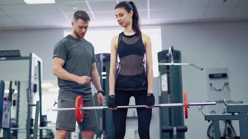 Woman Lifts Barbell with her Fitness Coach at Gym
