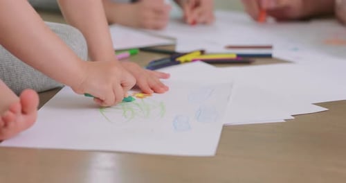 Nahaufnahme der Hände eines kleinen Mädchens, das mit Bleistiften auf dem weißen Papier auf dem Boden zeichnet