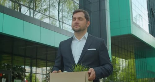 Un homme d'affaires désespéré a perdu son emploi et marche près d'un centre de bureaux avec une boîte en carton et des documents avec un