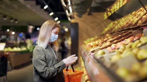 young blonde woman in a protective face medical mask picks chooses apple fruits vegetables