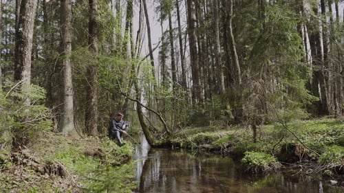 Einsamer Mann sitzt am überwucherten Flussufer im Wald führt Holzzweig ins Wasser