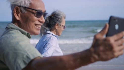 senior couple relaxing outdoors walking and taking selfie together with smartphone on beach