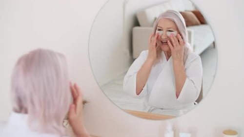 Senior Woman Applies Eye Patches in Bathroom Mirror