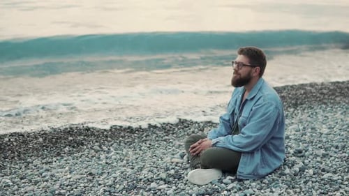 Ein junger und bärtiger Mann sitzt an einem steinigen Strand und genießt den Meerblick