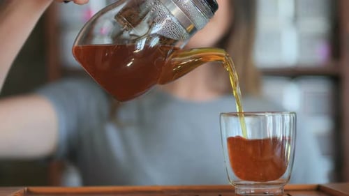 Weibliche Hand gießt roten Tee vom Glaskessel zur kleinen Tasse