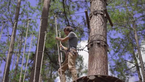 A Young Man in an Adventure Park. He Wears a Safety Harness. He Climbs on a High Rope Trail. Outdoor