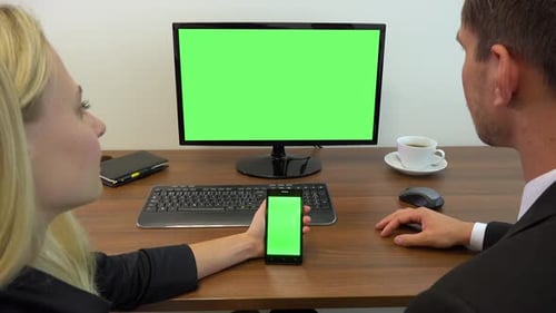 Two office workers sit at a desk and look at a computer and a smartphone with green screens