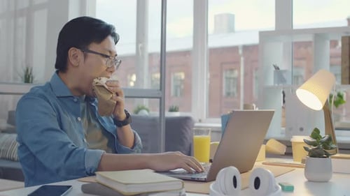 Asian Man Eating Sandwich and Using Laptop at Office Desk