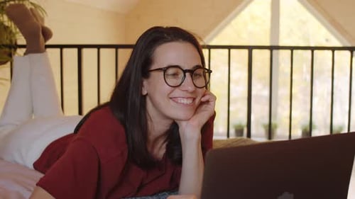 Fröhliche Frau liegt auf dem Bett und chattet im Web Call auf dem Laptop
