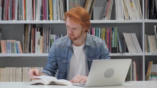 Rothaariger Mann liest Buch sitzend in Bibliothek