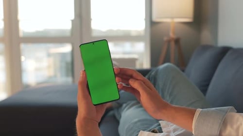 Man at Home Lying on a Sofa and Using Smartphone with Green Mock-up Screen in Vertical Mode. Guy