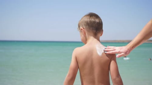 Mutter legt Sonnencreme auf den Rücken ihres Sohnes, während sie an einem sonnigen Tag an einem Sandstrand steht