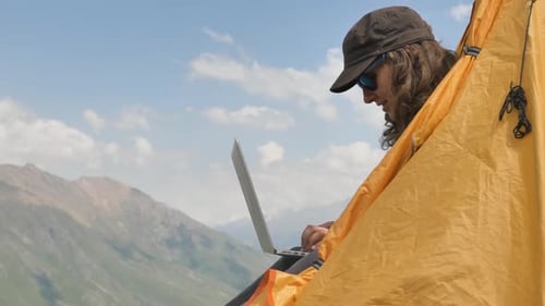 Senhora digita no laptop sentada em uma tenda amarela contra a encosta
