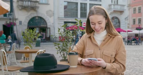 The Girl is Sitting at a Table in a Cafe and Texting on the Phone