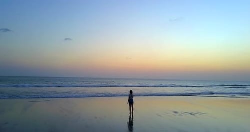 Drohnenaufnahme einer jungen Frau, die bei Sonnenuntergang am Strand spaziert.