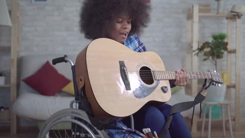 Disabled African American Woman with an Afro Hairstyle in a Wheelchair Plays Acoustic Guitar Close