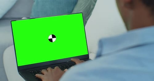 Mixed Race Ethnic Man Lying on Sofa at Home Uses Laptop with Green Mockup Screen