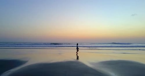 Aerial drone view of a young woman walking on the beach at sunset.