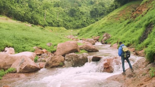 A young male trekker with backpack standing on the rock of a stream in a green valley.