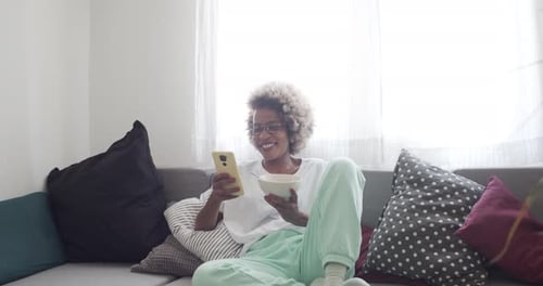 Woman Enjoys Cereal While Using Smartphone on Sofa