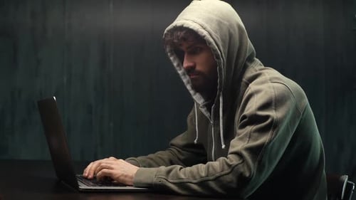 Seitenansicht eines gefährlichen konzentrierten jungen bärtigen Mannes, der die Online-Website am dunklen Arbeitsplatz hackt.