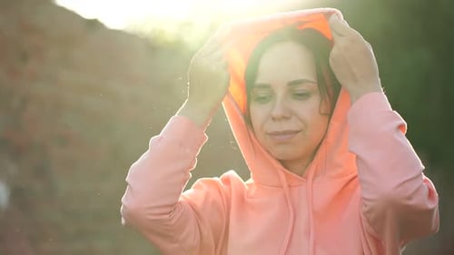 Portrait of Young Woman Putting on Hood on Street. Adult Female Puts Hood on Head in Cool Weather