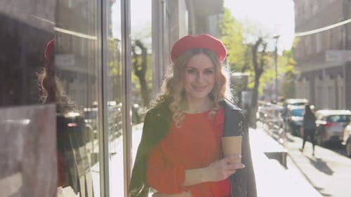 Stylish Young Blonde Woman in a Red Dress and Black Braid Stands on a Cozy Street in the Morning Sun