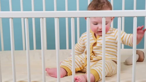 Infant baby boy is sitting in a crib and playing with toys, blue studio background. Caucasian toddle