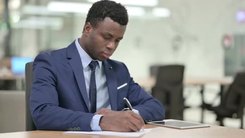 Hardworking African Businessman Writing on Paper in Office
