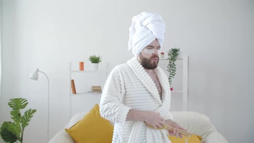 Sorgloser lustiger junger Mann, der Spaß daran hat, alleine zu Hause in einem Bademantel und einem Handtuch auf dem Kopf zu tanzen
