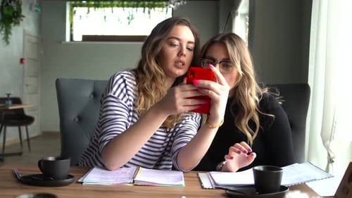 Young Cheerful Girls-best Friends Are Sitting in a Cafe, Using a Smartphone and Laughing
