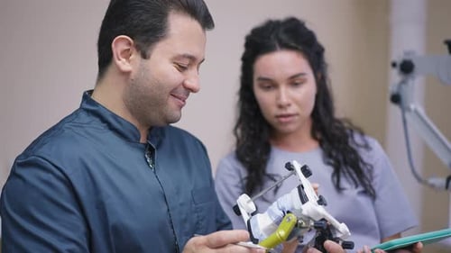 Side View Expert Male Orthodontist with Dental Cast and Blurred Curios Student Talking at Background