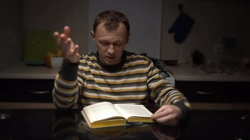 Young Man with Expression Reads a Book While Sitting at a Table in the Evening in the Kitchen