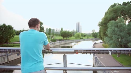 Ein Typ in einem blauen T-Shirt steht in einem Park auf einer Brücke und schaut auf den Fluss, Kamerabewegung