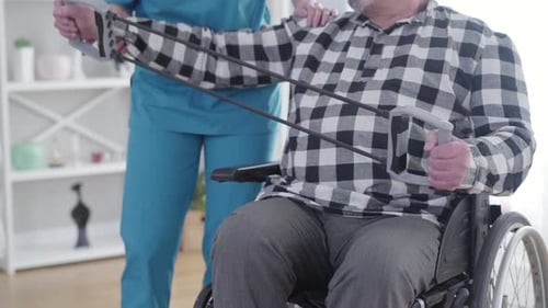 Unrecognizable Female Invalid Tender Stretching Elastic Band Together with Patient in Nursing Home