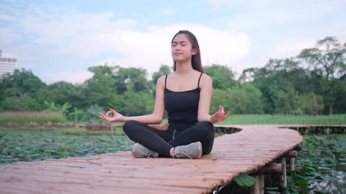 Asian sport woman sit on bamboo bridge with yoga posture near lotus pond in green park